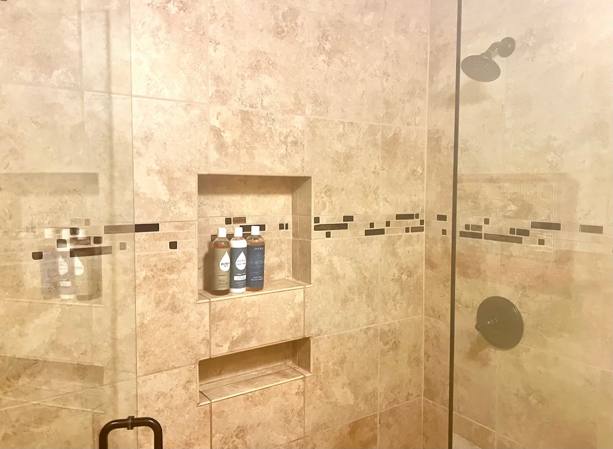A close-up view of a stylish shower area, highlighting a rainfall showerhead, tiled walls, organized bottles on a shelf, and a serene, spa-like ambiance. The neutral color palette adds to the calming effect.
