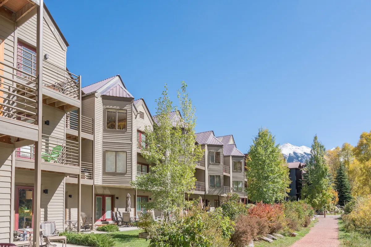 A picturesque scene of Viking Lodge’s multi-story buildings set against a lush green landscape and a stunning mountain backdrop. People are seen enjoying the outdoors, relaxing on balconies, and strolling along scenic garden pathways. The riverfront gardens and open areas invite both tranquility and community gatherings.