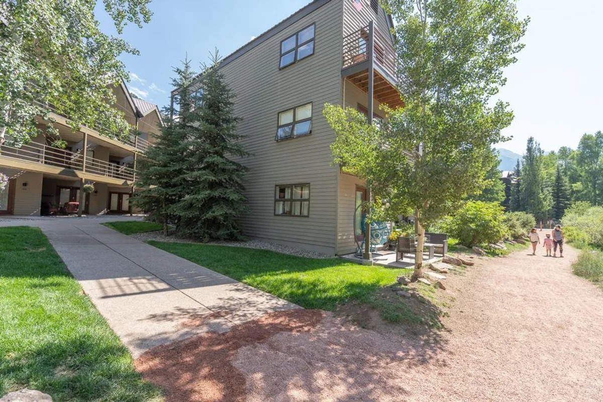 A welcoming exterior scene showing the building nestled among trees, a pathway in the foreground, and people walking and exploring the outdoor space. The area feels lived-in and friendly, with greenery and a bright, sunny atmosphere.