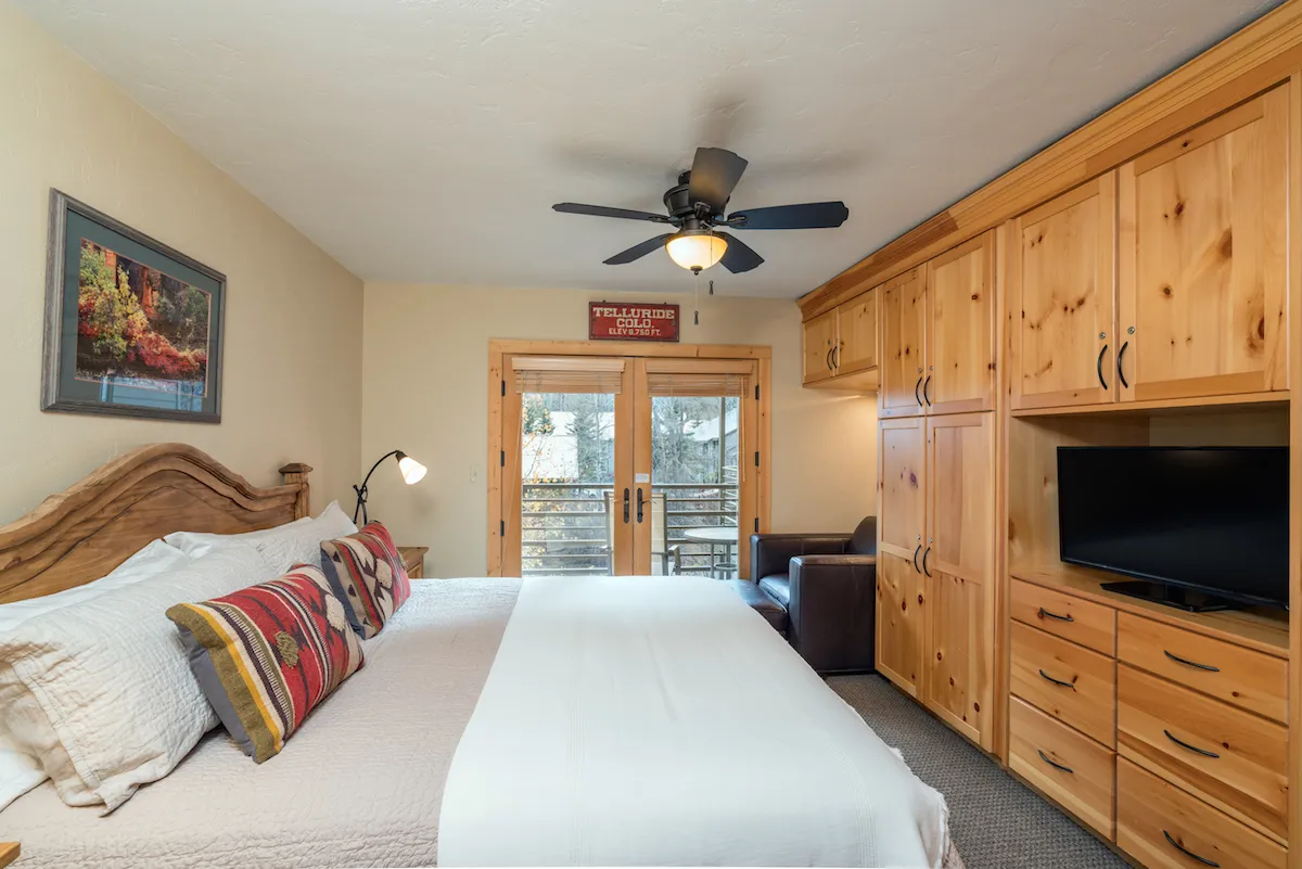 The image shows a cozy bedroom interior with a comfortable bed adorned with decorative pillows, set near sliding glass doors that fill the space with natural light. Wooden furniture and tasteful framed artwork add rustic charm, while the doors open to a balcony that hints at mountain views—perfect for relaxing or reading a book while enjoying the Colorado scenery.