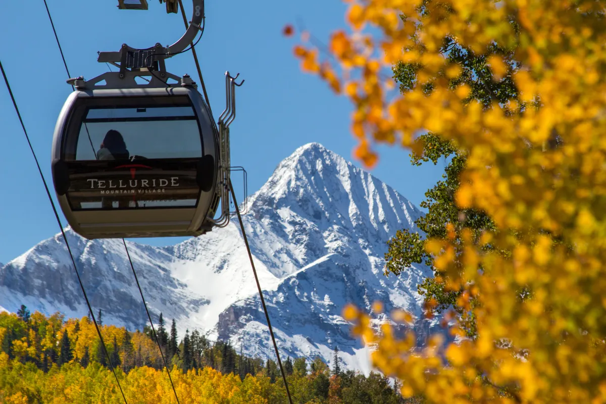 A gondola glides above a landscape combining golden autumn foliage and snow-capped mountains. The scene exudes peaceful adventure, with the promise of both winter sports and scenic sightseeing rides.