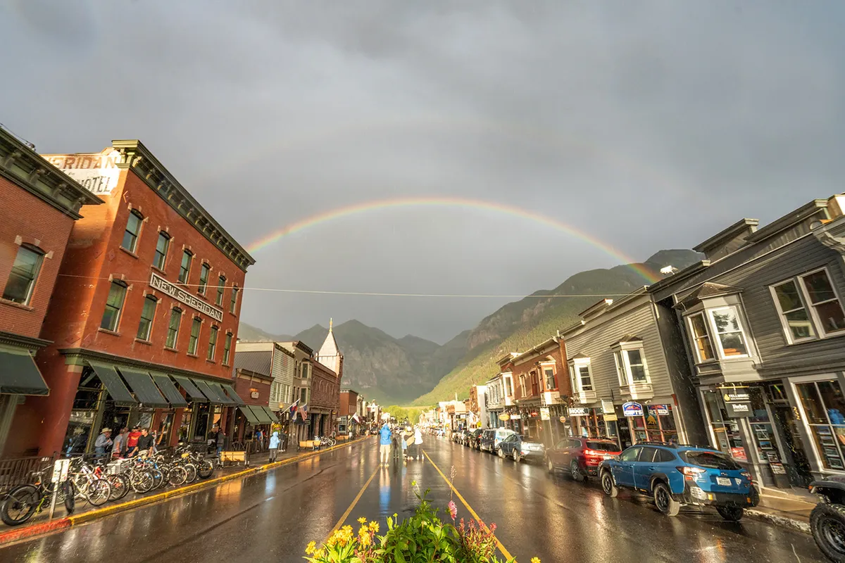 A vibrant downtown street scene in Telluride featuring historic buildings glistening with rain reflections, a rainbow arching overhead, several bicycles parked, and people walking and shopping. The street feels alive and welcoming, capturing the essence of a walkable, engaging town center.