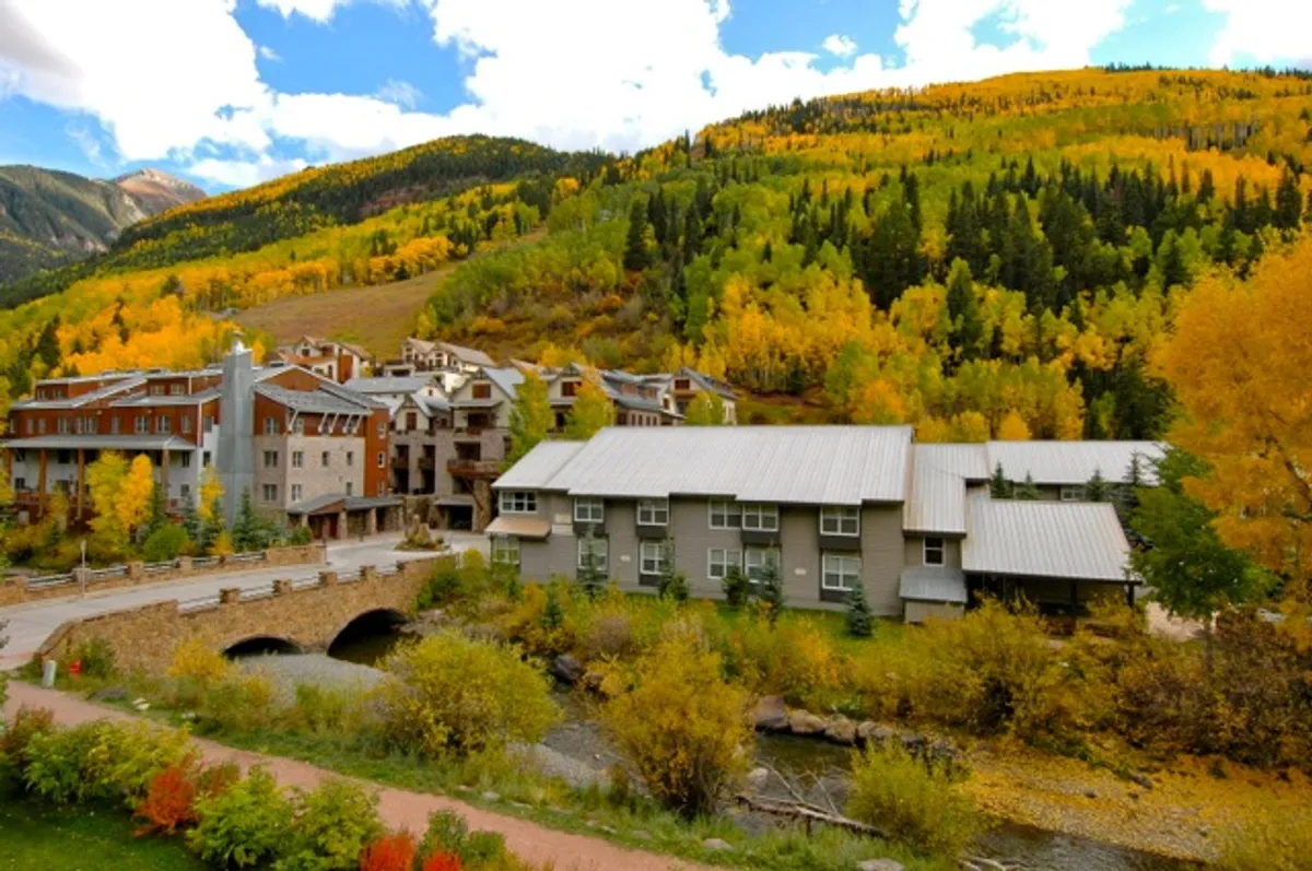 A serene riverside setting displays colorful autumn foliage, contemporary architecture with balconies, and the calm river reflecting the mountain backdrop. The scene invites romantic strolls, relaxation on private balconies, and quiet connection with nature.
