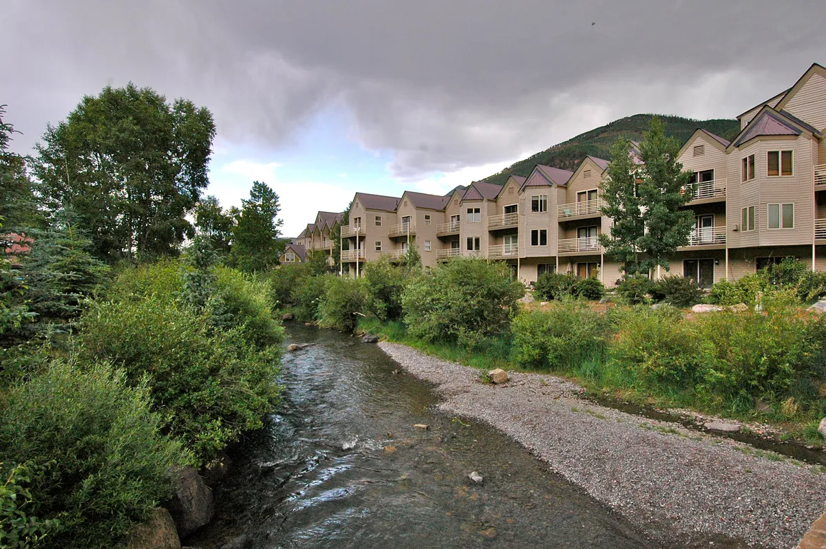A peaceful exterior shot that frames the stream in the foreground, bordered by lush green trees, with the condo buildings set back from the water. A pathway meanders alongside the stream, inviting families for walks or wildlife watching. The scene is serene and inviting, suggesting easy access to nature right outside the property.