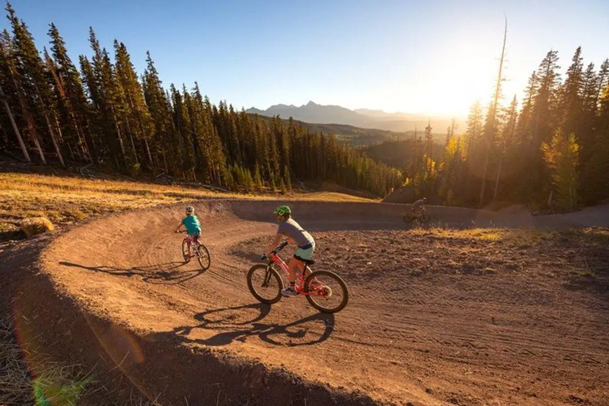 This exterior image features mountain bike riders navigating a winding trail surrounded by tall trees, with a dramatic sunset sky overhead. The scene is energetic and action-packed, highlighting the thrill of mountain biking and hiking in Telluride's natural landscape.