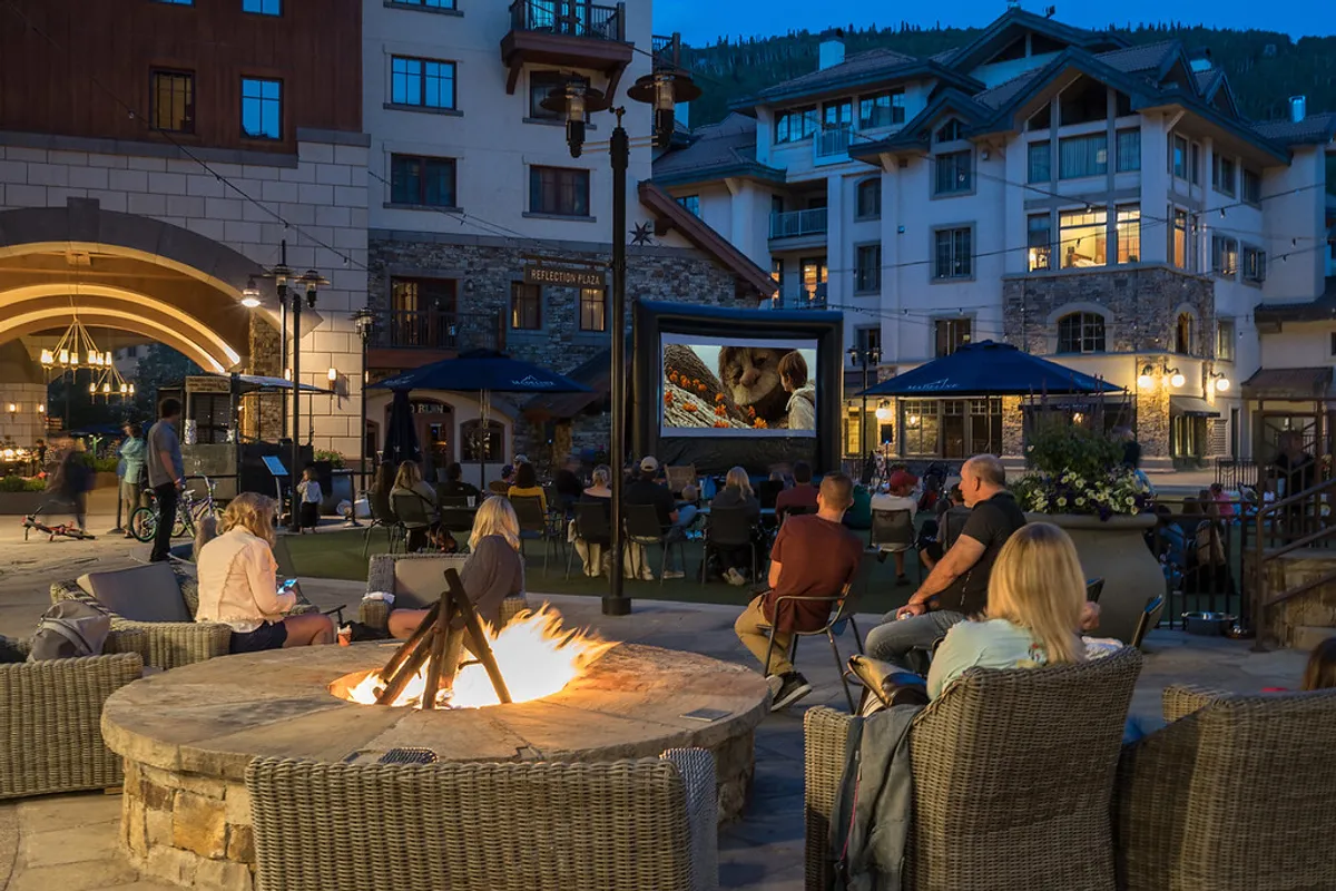 A communal outdoor area featuring a fire pit, movie screen, comfortable seating, and surrounding buildings. People are gathered around, socializing and enjoying the open-air setting, which is both accessible and central.