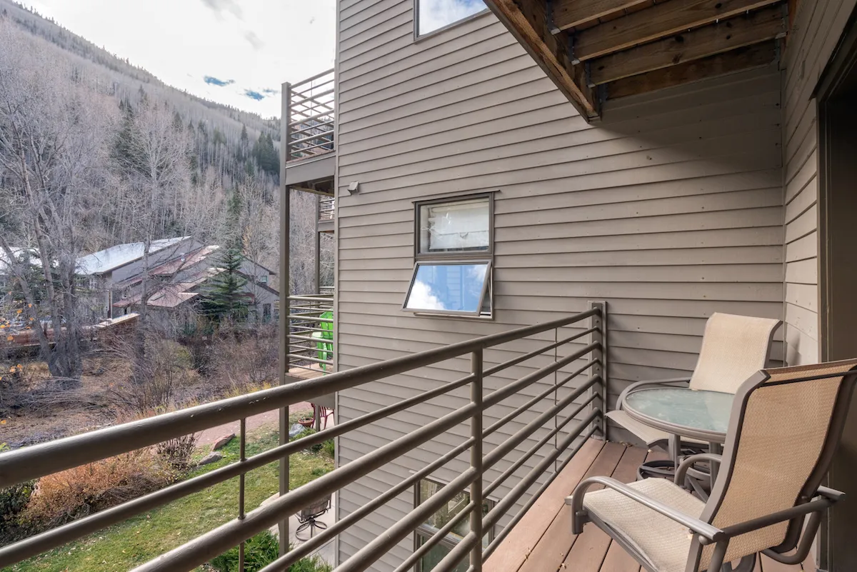 A private balcony scene featuring a sturdy railing, a table and chairs, and views of surrounding trees and distant mountains. The tranquil setting suggests enjoying morning coffee, reading, or quietly appreciating the natural beauty and peaceful atmosphere.