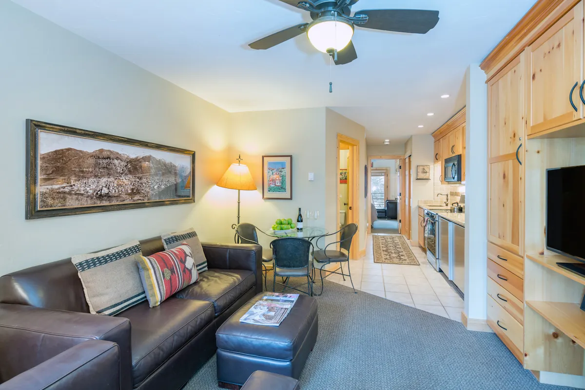 A spacious open-plan living area featuring a plush sofa with decorative pillows, a dining table with chairs, tasteful wall art, a lamp adding a warm glow, and a view into the adjacent kitchen area. The room is arranged for both relaxation and socializing, with natural light enhancing the inviting ambiance.