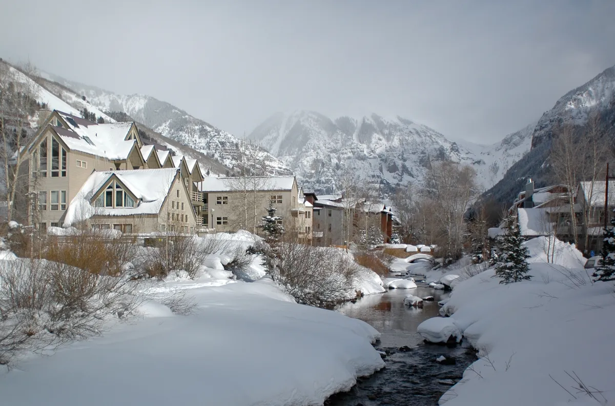 A scenic winter scene with snow-covered mountains rising above a gently flowing river bordered by snow-laden trees. The architecture of the nearby buildings hints at cozy accommodations nestled in the landscape, with the tranquil ambiance suggesting a perfect romantic retreat after a day of skiing or exploring.